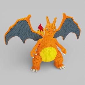 Könnte beinhalten: Handgefertigte Charizard-Figur aus orangefarbenem und gelbem Häkelgarn mit grauen Flügeln und einer roten Flamme. Die Figur hat weiße Krallen und steht auf einer grauen Oberfläche. Die Flügel sind ausgebreitet.