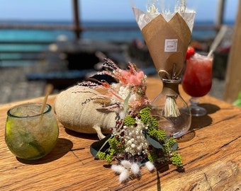 Può includere: Due cocktail, uno con una cannuccia, su un tavolo di legno con un bouquet di fiori secchi in un vaso di vetro. I fiori sono in un involucro di carta marrone con la parola "ALMA" stampata su di esso. Il tavolo è all'aperto con vista sull'oceano sullo sfondo.