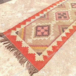 May include: A long, rectangular rug with a geometric pattern in shades of brown, pink, and orange. The rug has a fringe border and is made of a woven material.