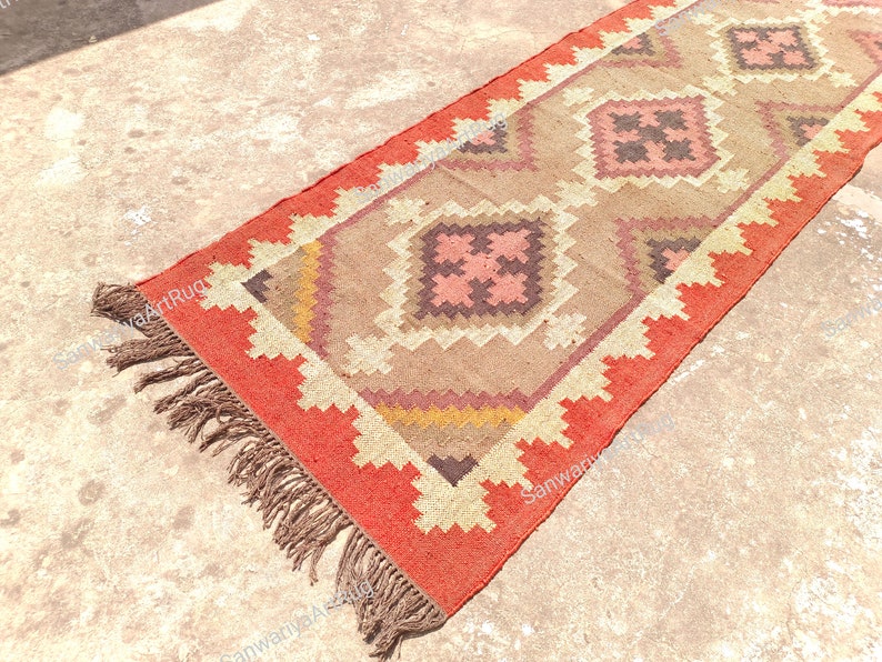 May include: A long, rectangular rug with a geometric pattern in shades of brown, pink, and orange. The rug has a fringe border and is made of a woven material.