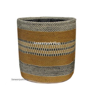 May include: A cylindrical woven basket with a textured design. The basket features horizontal bands of tan, black, and white. The text "SanwariyaArtRug" is visible on the side. The basket is suitable for storage or decorative purposes.