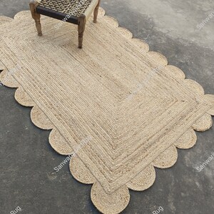 May include: A natural fiber rug with a scalloped edge. The rug is made of jute and has a light brown color. The rug is placed on a concrete floor and a wooden chair is sitting on top of it.