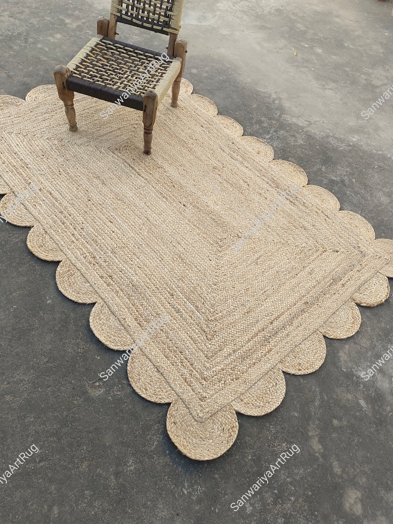 May include: A natural fiber rug with a scalloped edge. The rug is made of jute and has a light brown color. The rug is placed on a concrete floor and a wooden chair is sitting on top of it.