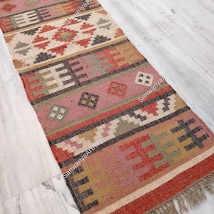 Puede incluir: Una alfombra rectangular larga con un patr&oacute;n geom&eacute;trico en tonos rojos, marrones, beige y rosa. La alfombra tiene flecos en el borde inferior.