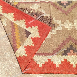May include: A red, brown, and white geometric patterned rug with fringe. The rug is folded in half and the fringe is visible on the bottom edge.