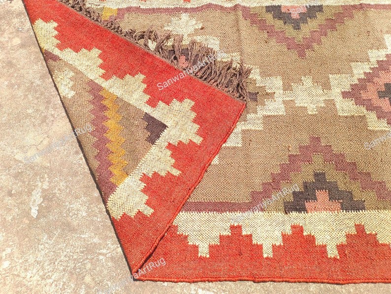 May include: A red, brown, and white geometric patterned rug with fringe. The rug is folded in half and the fringe is visible on the bottom edge.