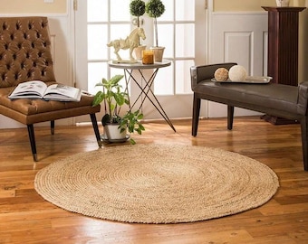 Puede incluir: Una alfombra redonda de yute natural de color beige claro con textura tejida. La alfombra está colocada sobre un suelo de madera dura en un salón con una silla de cuero marrón y un banco.