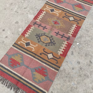 May include: A long, narrow rug with a geometric pattern in shades of red, brown, gray, and beige. The rug has a fringe on both ends.