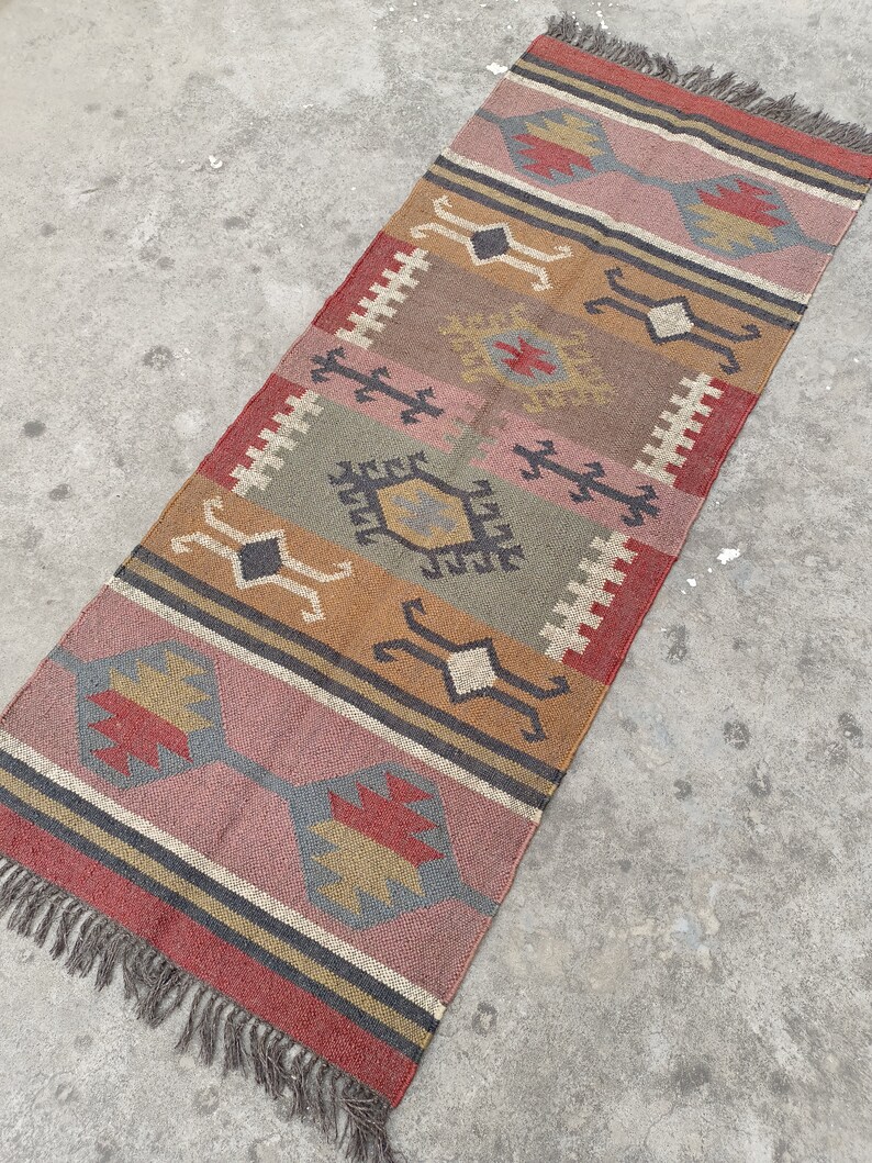 May include: A long, narrow rug with a geometric pattern in shades of red, brown, gray, and beige. The rug has a fringe on both ends.