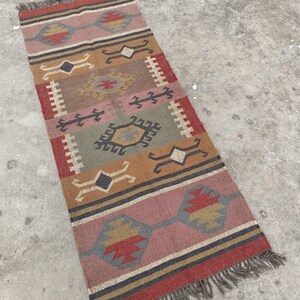 May include: A colorful, geometric patterned rug with fringe on the ends. The rug is made of jute and has a variety of colors, including red, orange, yellow, green, and brown.