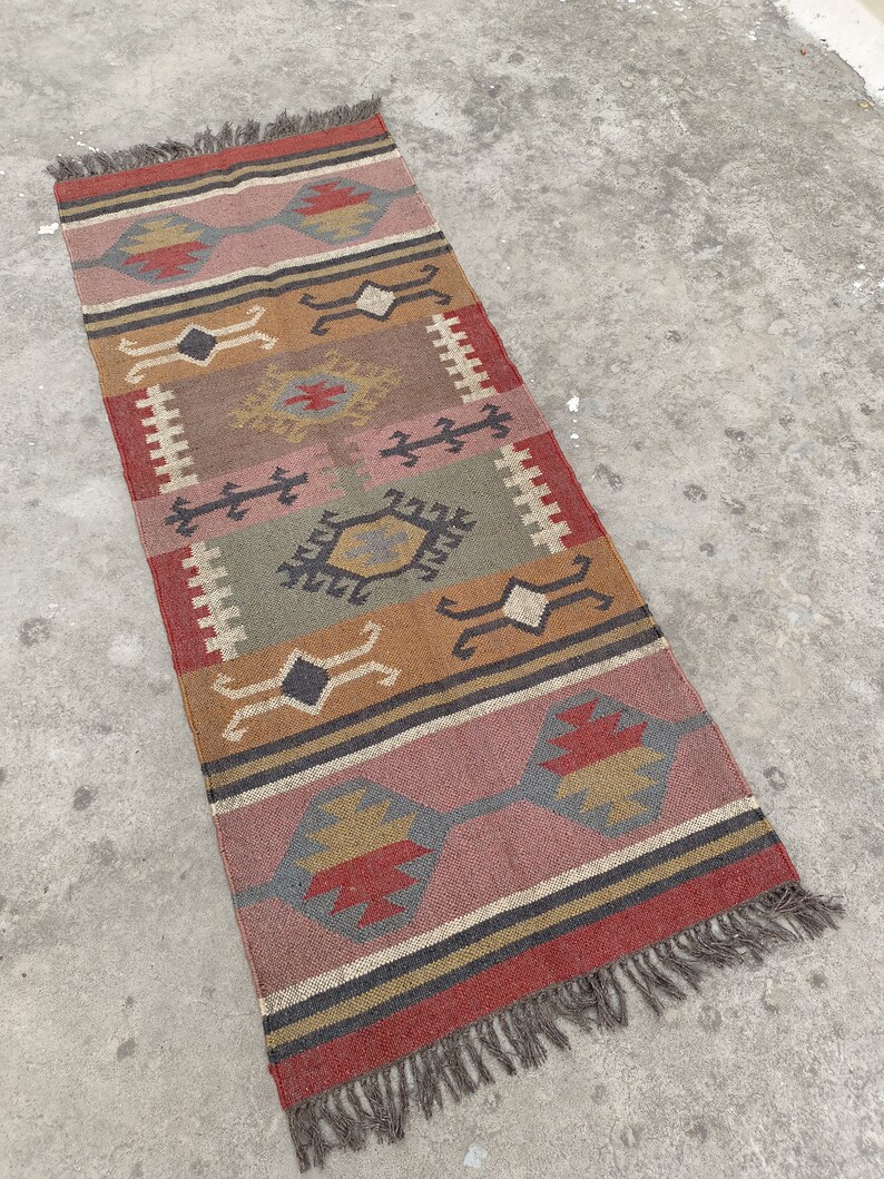 May include: A colorful, geometric patterned rug with fringe on the ends. The rug is made of jute and has a variety of colors, including red, orange, yellow, green, and brown.