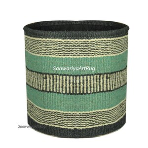 May include: Cylindrical woven basket with horizontal stripes in teal, black, and cream. The basket has a dark gray rim and base. The text "SanwariyaArtRug" is printed on the side of the basket.