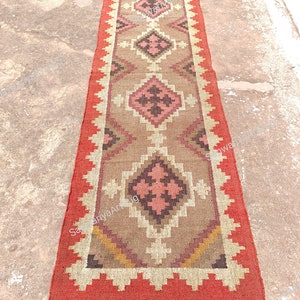 May include: A long, rectangular rug with a geometric pattern in shades of brown, pink, and white. The rug has a red border and brown fringe.
