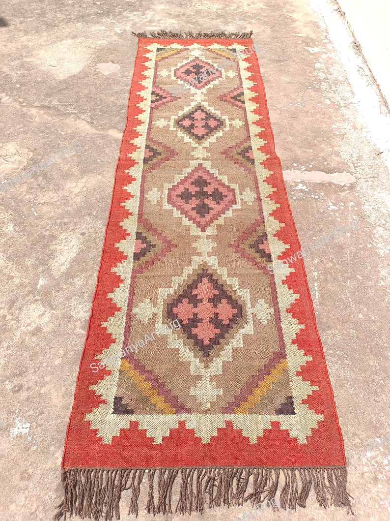 May include: A long, rectangular rug with a geometric pattern in shades of brown, pink, and white. The rug has a red border and brown fringe.