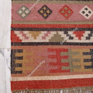Puede incluir: Una alfombra tejida a mano colorida con patrones geom&eacute;tricos en tonos rojos, rosas, marrones y verdes. La alfombra tiene un borde de flecos y est&aacute; hecha de fibras naturales.