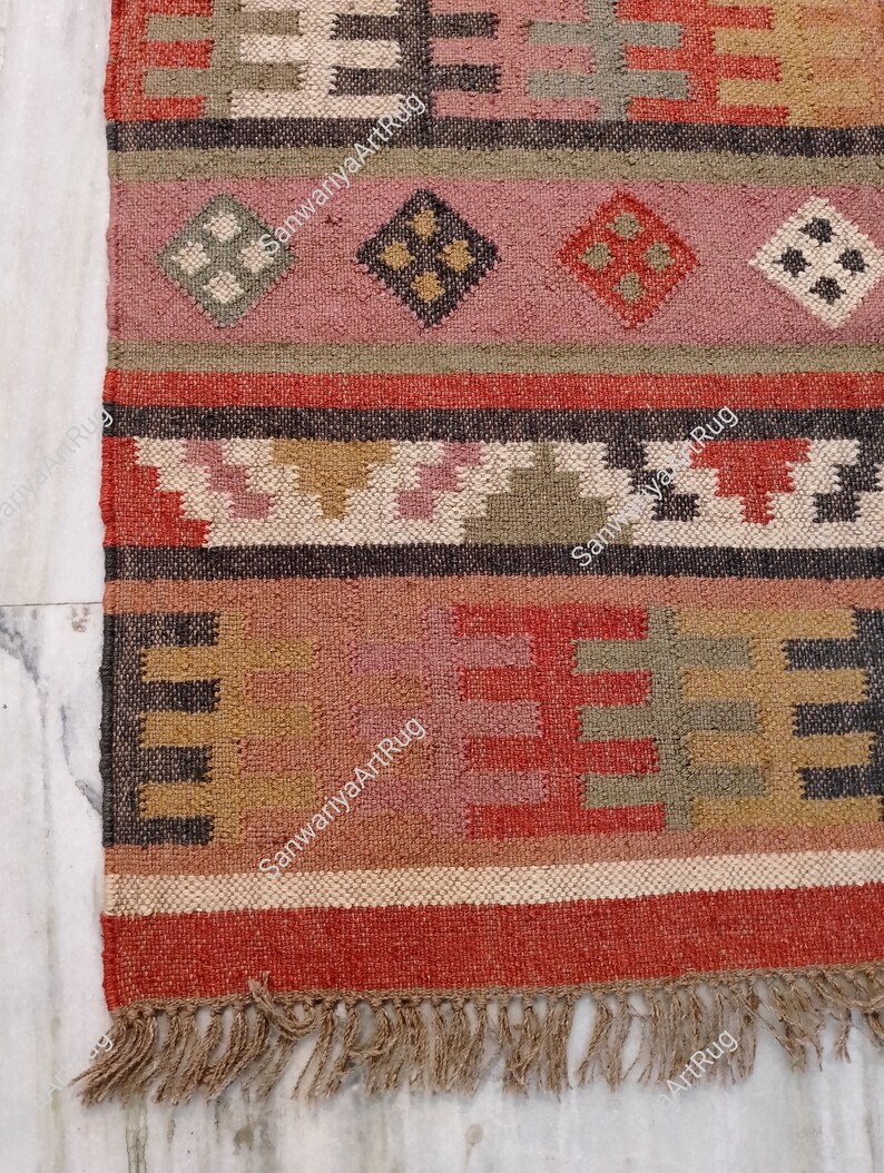 Puede incluir: Una alfombra tejida a mano colorida con patrones geom&eacute;tricos en tonos rojos, rosas, marrones y verdes. La alfombra tiene un borde de flecos y est&aacute; hecha de fibras naturales.