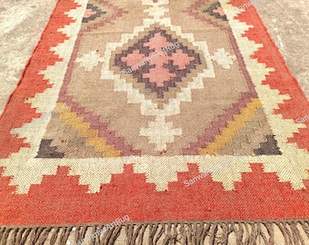 May include: A rectangular rug with a geometric pattern in shades of red, brown, beige, and pink. The rug features a central diamond design and a fringed edge. The border is a vibrant red with a zig-zag pattern.