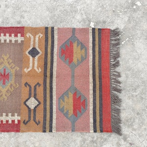 May include: A colorful rug with geometric patterns in shades of red, brown, gray, and beige. The rug has a fringe on the right side.