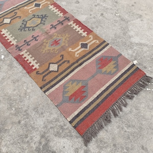 May include: A long, rectangular rug with a colorful geometric pattern in shades of red, orange, brown, gray, and pink. The rug has a fringe on both ends.