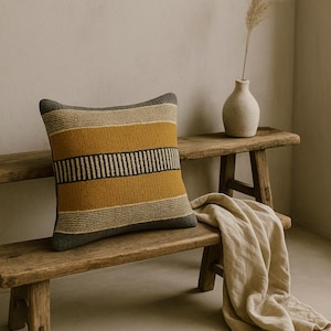 以下が含まれることがあります： 織り模様の正方形の装飾用クッション。マスタードイエロー、グレー、ブラックの横縞模様です。素朴な木製のベンチの上に置かれ、近くにはニュートラルカラーの毛布が掛けられています。落ち着いた雰囲気の画像です。