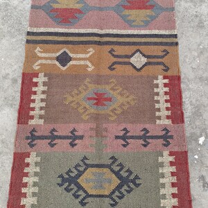 May include: A colorful, handwoven rug with a geometric pattern in shades of brown, red, pink, and gray. The rug has a fringe on the top and bottom.