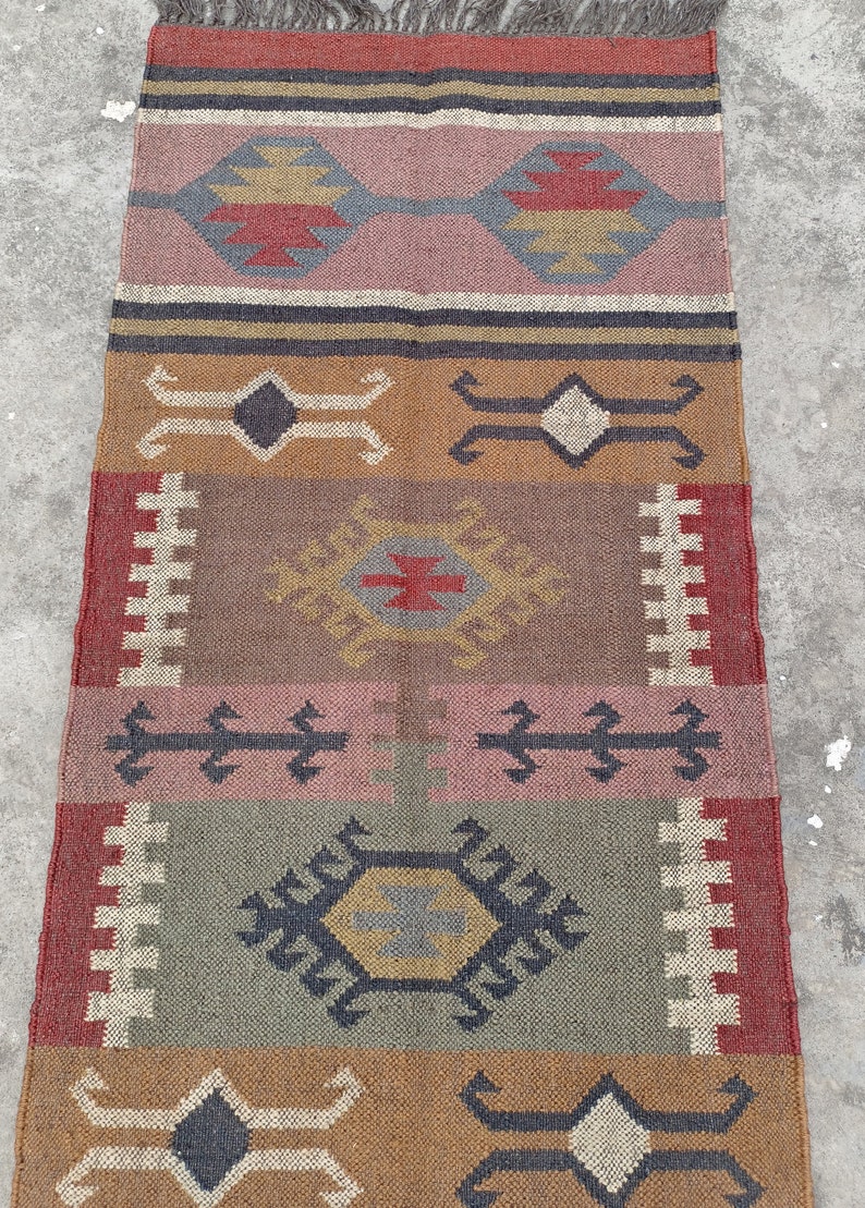 May include: A colorful, handwoven rug with a geometric pattern in shades of brown, red, pink, and gray. The rug has a fringe on the top and bottom.