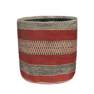 May include: A cylindrical woven basket with a red, black, and beige striped pattern. The basket has a black interior and a beige base. The woven texture is visible throughout the design, creating a rustic aesthetic.