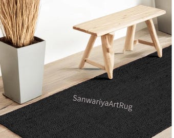 Op de afbeelding: Zwart geweven tapijt met een textuur. Het tapijt ligt op een houten vloer en een houten bank staat op de achtergrond.