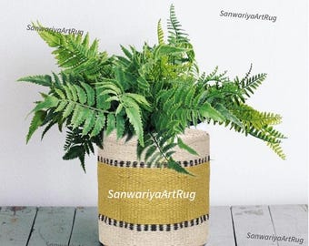 May include: A cylindrical woven basket with a yellow band and black stripes, filled with green fern plants. The basket is made of natural fibers and sits on a wooden surface. The text "SanwariyaArtRug" is visible on the basket.