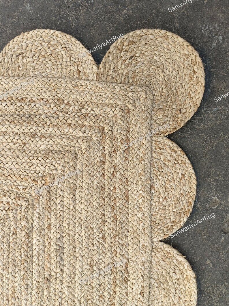 May include: A natural fiber rug with a flower shape. The rug is made of woven jute and has a light brown color. The rug has five rounded petals and a square center.