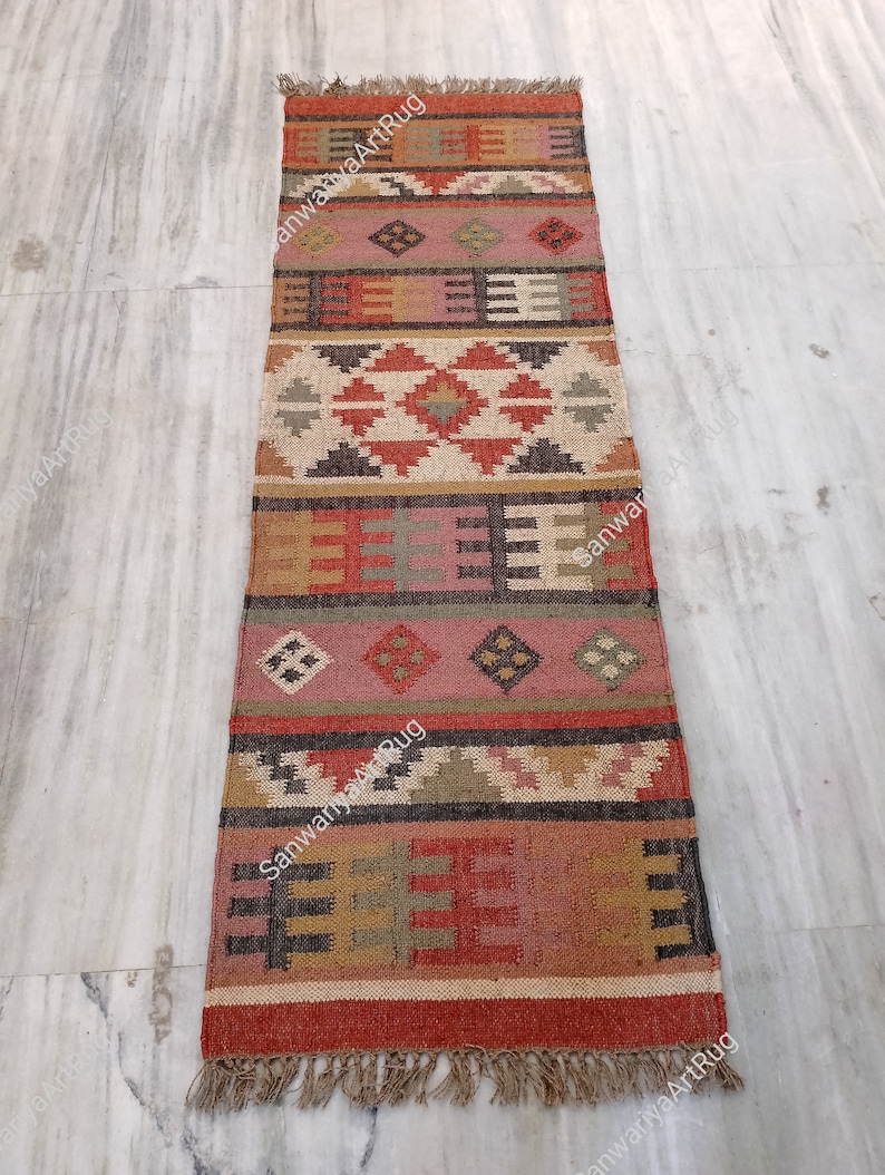 Puede incluir: Una alfombra de yute rectangular larga con un patr&oacute;n geom&eacute;trico en tonos de rojo, marr&oacute;n, beige y gris. La alfombra tiene flecos en ambos extremos.