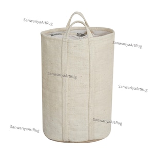 Puede incluir: Una cesta de almacenamiento cilíndrica grande de color blanco roto con textura tejida y base marrón. La cesta tiene dos asas y está hecha de fibras naturales.