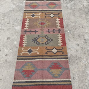 May include: A long, rectangular rug with a geometric pattern in shades of red, brown, green, and beige. The rug has a fringe on both ends.