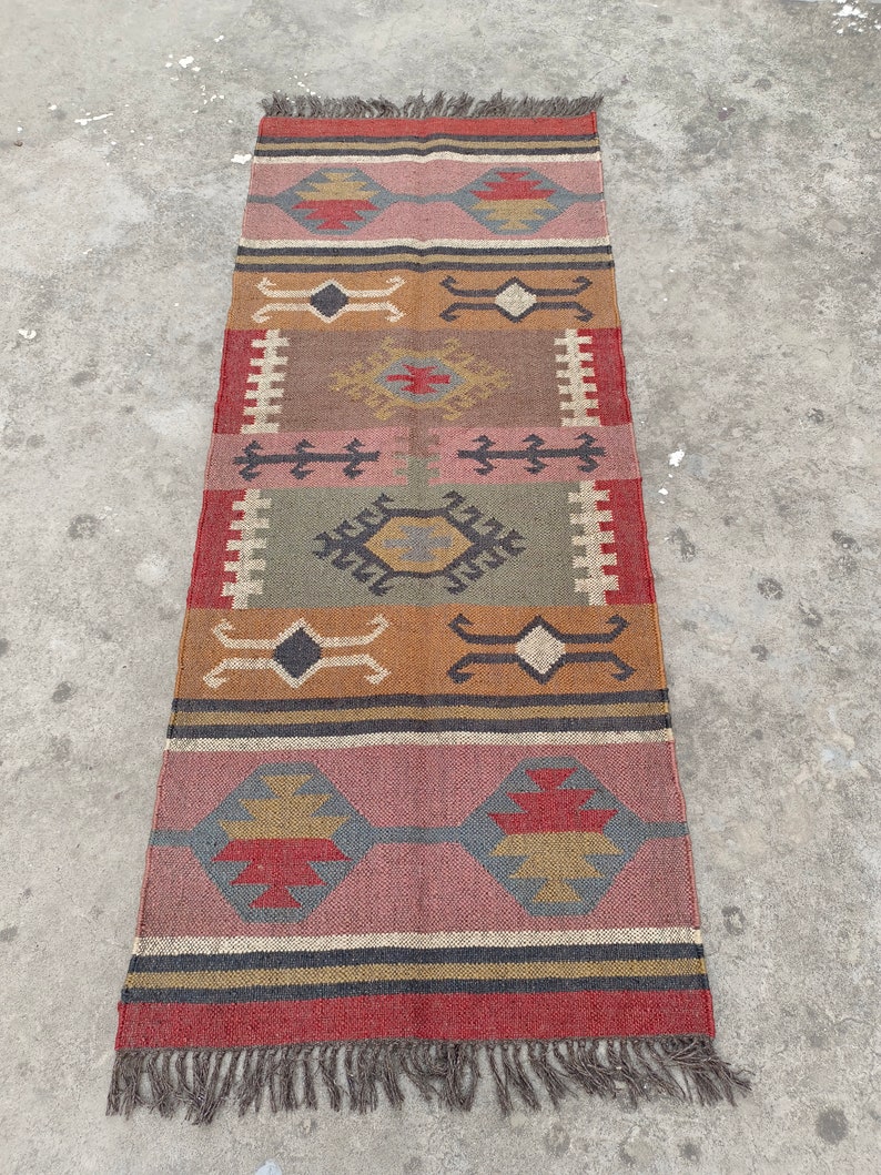 May include: A long, rectangular rug with a geometric pattern in shades of red, brown, green, and beige. The rug has a fringe on both ends.