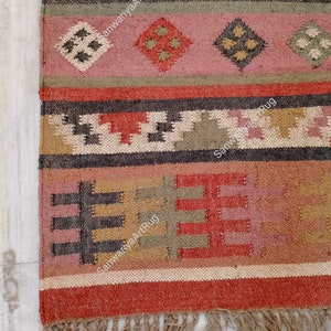 Puede incluir: Una alfombra tejida a mano colorida con patrones geom&eacute;tricos en tonos de rojo, marr&oacute;n, beige y verde. La alfombra tiene un borde de flecos y el texto "SanwariyaArtRug" est&aacute; tejido en la alfombra.