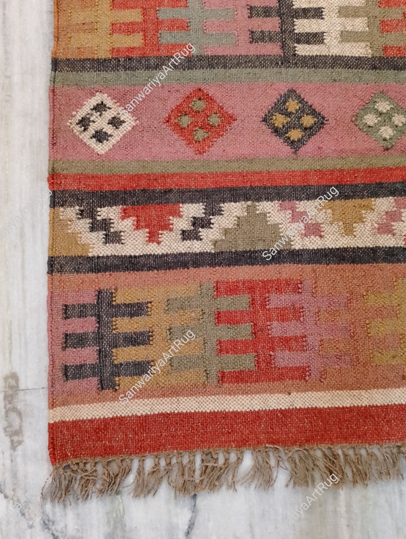 Puede incluir: Una alfombra tejida a mano colorida con patrones geom&eacute;tricos en tonos de rojo, marr&oacute;n, beige y verde. La alfombra tiene un borde de flecos y el texto "SanwariyaArtRug" est&aacute; tejido en la alfombra.