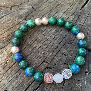 May include: A green and blue gemstone bracelet with silver beads and two silver tree of life charms. The bracelet is on a wooden surface.