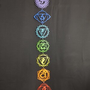 Op de afbeelding: Een wandhanger met zeven chakra's, waarbij elke chakra wordt weergegeven door een andere kleur en symbool. De chakra's zijn paars, indigo, blauw, groen, geel, oranje en rood.