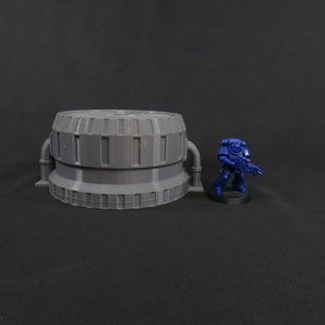 Könnte beinhalten: Ein graues, zylindrisches, 3D-gedrucktes Modell eines futuristischen Gebäudes mit Rohren und einer strukturierten Oberfläche. Das Modell ist etwa 10 cm hoch und steht neben einer blauen Plastikminiatur eines Space Marines.