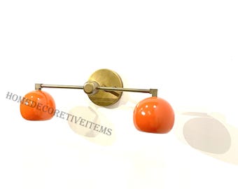 May include: A brass wall sconce with two orange globe-shaped light fixtures. The sconce has a simple, modern design.