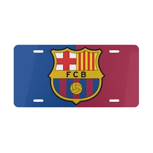 May include: A blue and maroon license plate with the FC Barcelona logo. The logo is a shield with a red cross, yellow and red stripes, and a soccer ball.