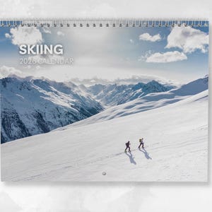 May include: A spiral-bound 2026 calendar featuring a snowy mountain landscape with two skiers. The calendar cover displays the word "SKIING" above "2026 CALENDAR" in a light gray font. The background shows a bright blue sky with clouds.