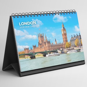Könnte beinhalten: Ein Tischkalender mit einem Foto von London, das das Parlament und den Big Ben unter blauem Himmel zeigt. Der Kalender hat einen schwarzen Ständer und eine Spiralbindung. Der Text "LONDON 2026 DESK CALENDAR" ist zu sehen.