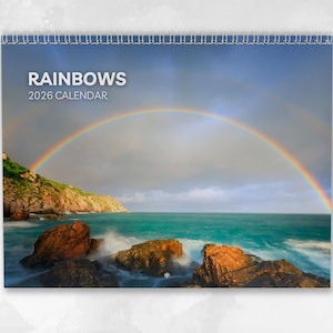 Könnte beinhalten: Ein Spiral-Kalender 2026 mit einem lebendigen Doppelregenbogen über einem türkisfarbenen Meer und einer felsigen Küste. Der Kalenderumschlag zeigt den Titel "RAINBOWS" in Weiß, darunter "2026 CALENDAR".