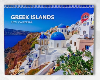 May include: A 2027 wall calendar titled GREEK ISLANDS featuring a photograph of white buildings with blue domes overlooking a deep blue sea and rocky cliffs. The calendar is bound with a silver wire spiral at the top edge against a white background.