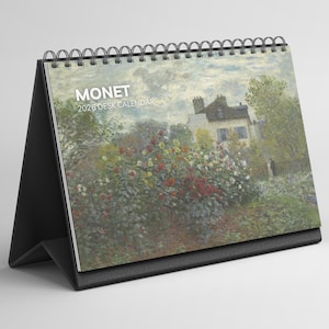 Puede incluir: Un calendario de escritorio con una pintura de Monet de un jardín y una casa. El calendario tiene un soporte negro y una encuadernación en espiral. El texto "MONET 2026 DESK CALENDAR" está impreso en la parte delantera.