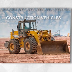 May include: A 2027 wall calendar titled CONSTRUCTION VEHICLES featuring a large yellow front-end loader parked on a dirt construction site under a cloudy blue sky. The calendar is bound with a silver wire spiral at the top edge.