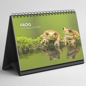 Op de afbeelding: Een bureaukalender met een foto van twee kikkers op een mosachtige tak. De kalender is voorzien van de tekst "FROG 2026 DESK CALENDAR". Hij heeft een zwarte standaard en een spiraalbinding.