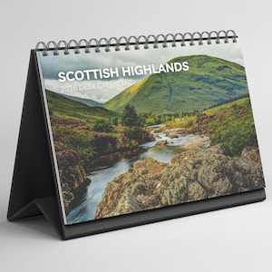 May include: A desk calendar with a black stand, spiral binding, and a scenic image of the Scottish Highlands. The cover reads "SCOTTISH HIGHLANDS 2026 DESK CALENDAR" above a landscape of mountains and a river, evoking a sense of nature.