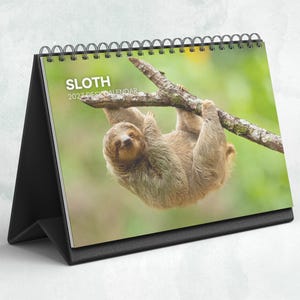 May include: A desk calendar with a black stand and a spiral binding at the top. The cover features a photograph of a brown sloth hanging from a tree branch against a green background. The text SLOTH 2027 DESK CALENDAR is printed in white.